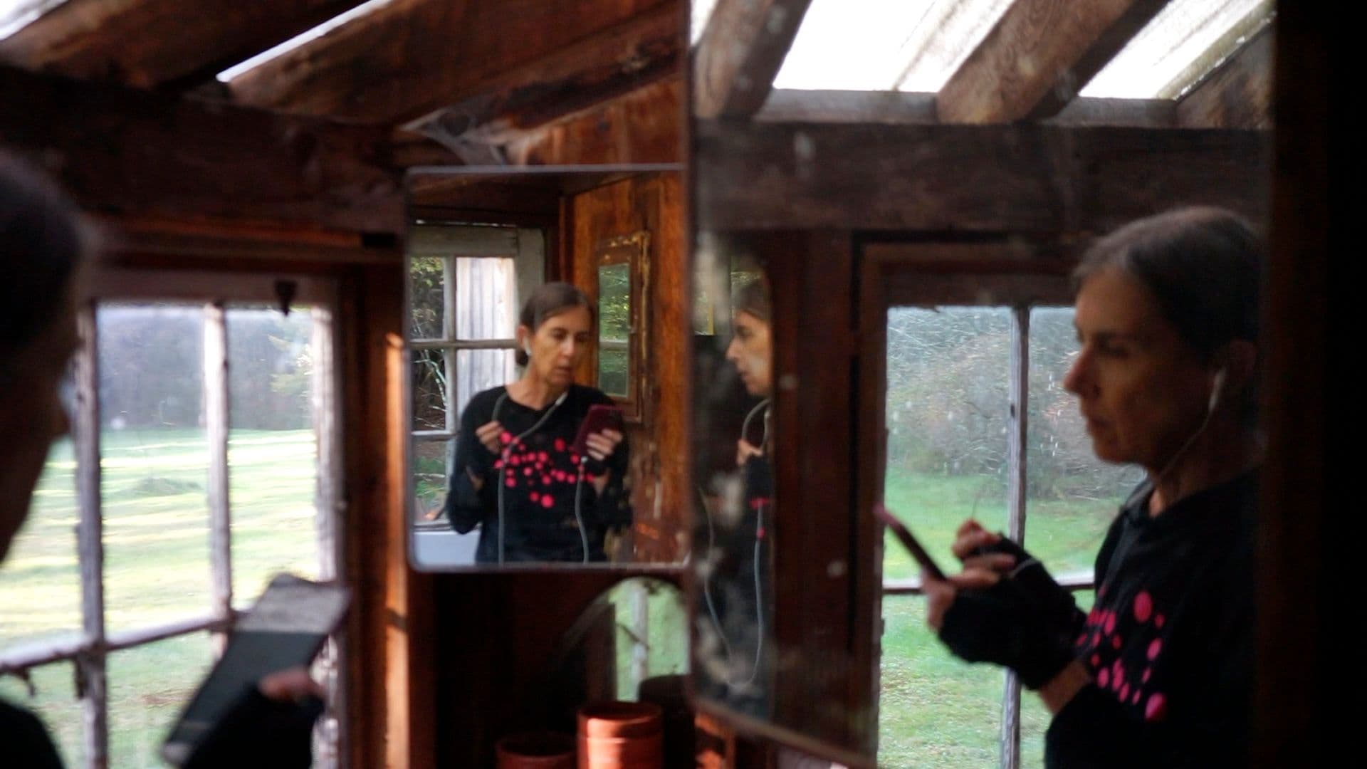 A woman is looking at herself in in multiple mirrors while speaking into corded headphones.