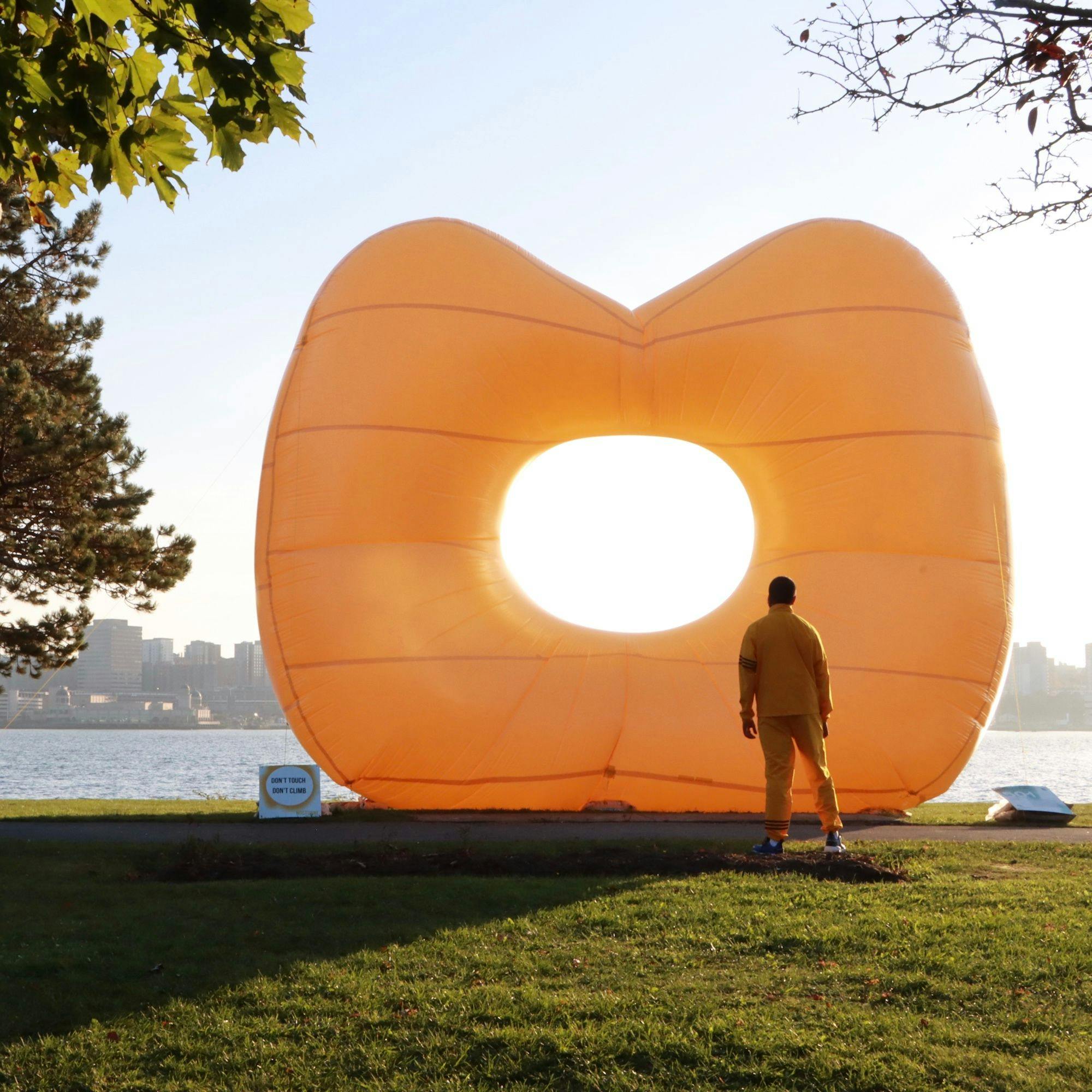A man standing near a waterfront in front of a large orange balloon with a hole in the middle.