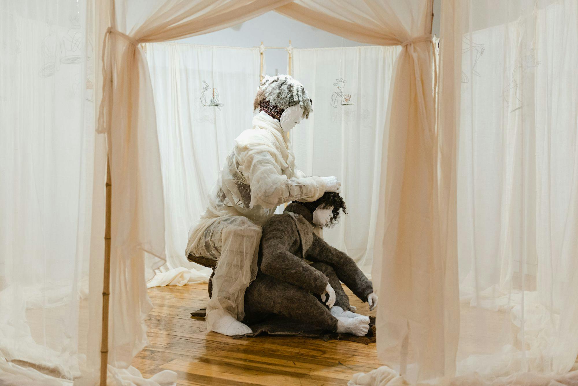 Two masked people crouch in a room covered with sheer white curtains. One figure crouches over the other with their hands in the other person’s hair. 