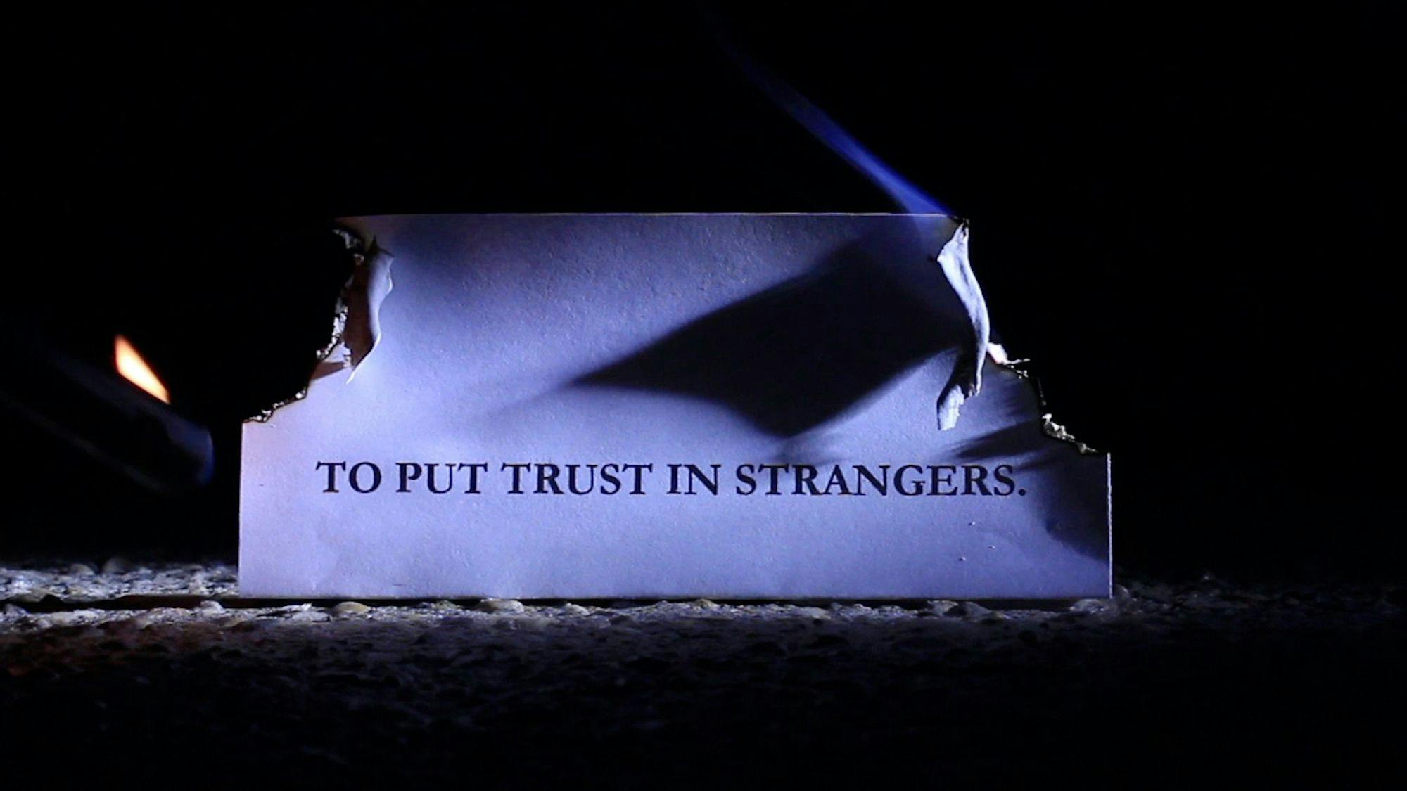 A piece of paper with the words “TO PUT TRUST IN STRANGERS” burning at the corners.  